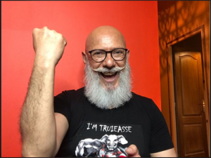 Photo de Juan Carlos de La Fistinière levant le poing, souriant, avec une barbe blanche et des lunettes.